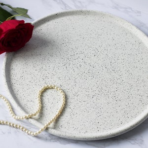 May include: A white ceramic tray with a speckled finish. The tray is round and has a slightly raised edge. A string of white pearls is draped over the tray.