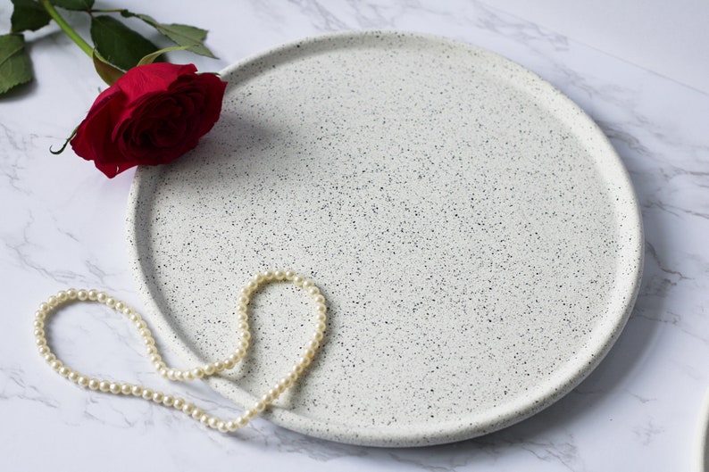 May include: A white ceramic tray with a speckled finish. The tray is round and has a slightly raised edge. A string of white pearls is draped over the tray.