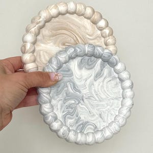 May include: Two decorative coasters. One is a light beige with a marbled design, and the other is gray and white with a similar pattern. Both have a beaded edge.