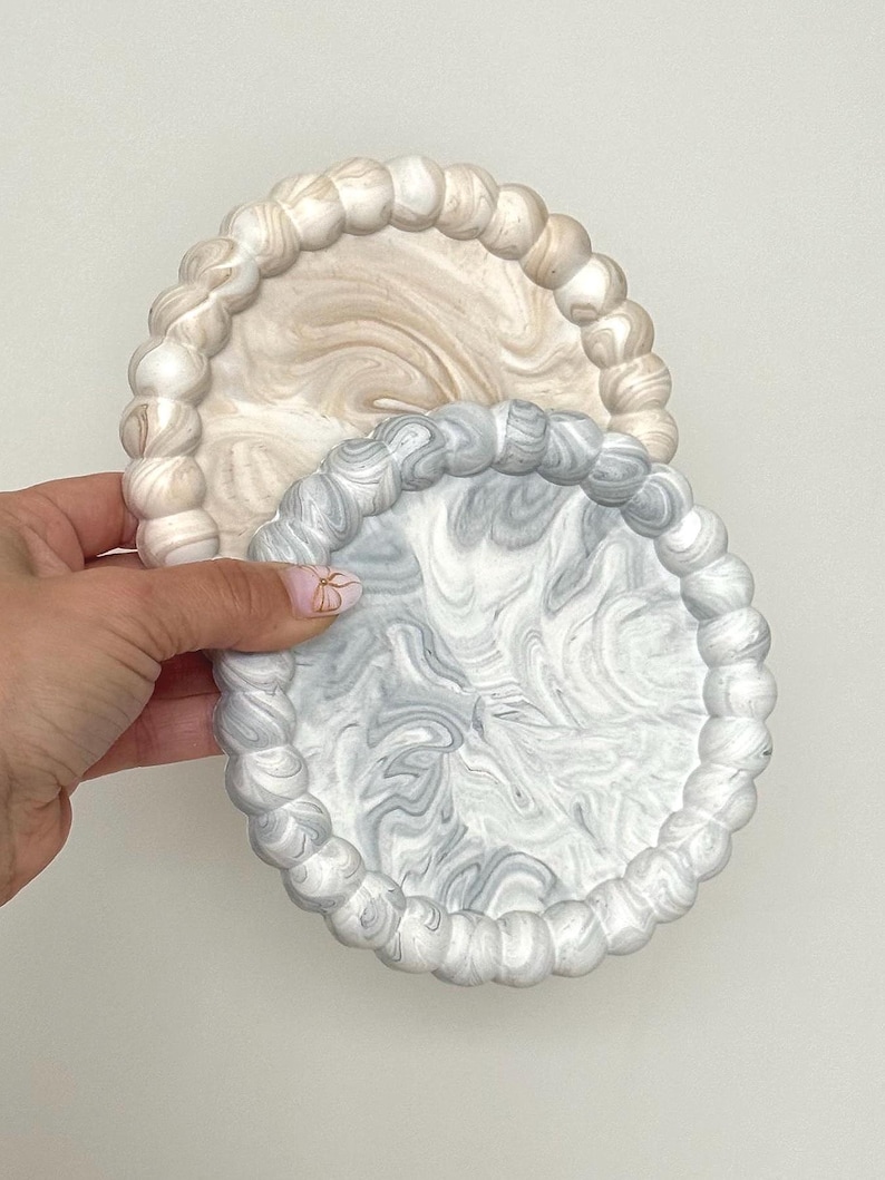 May include: Two decorative coasters. One is a light beige with a marbled design, and the other is gray and white with a similar pattern. Both have a beaded edge.
