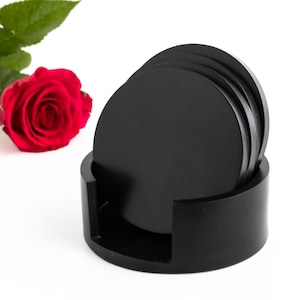 Pode incluir: Um conjunto de  copos de ardósia preta com um suporte de ardósia preta correspondente. Os copos são redondos e têm uma superfície lisa e polida.