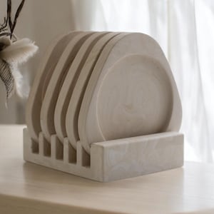 Pode incluir: Um conjunto de seis bases para copos bege, com formato quadrado e cantos arredondados, exibidas em um suporte correspondente. As bases e o suporte têm uma aparência marmorizada, sugerindo um material de pedra ou resina. O conjunto está em uma superfície de madeira clara.