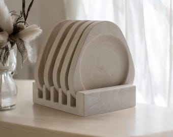 May include: A set of six beige, square-shaped coasters with rounded corners, displayed in a matching holder. The coasters and holder have a marbled appearance, suggesting a stone or resin material. The set is on a light-colored wooden surface.