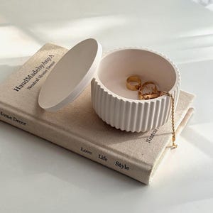 以下が含まれることがあります： クリーム色のリブ付きジュエリーボックス。蓋を開けると、金色の指輪とネックレスが見える。ボックスは「HandMadebyAnyA」と「Neutral Home Decor」の文字が書かれた本の上に置かれている。本には「Love Life Style」の文字もある。