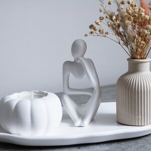 May include: White ceramic tray with a white pumpkin shaped candle, a white ceramic sculpture of a person sitting with their chin on their hand, and a white ceramic vase with dried flowers.
