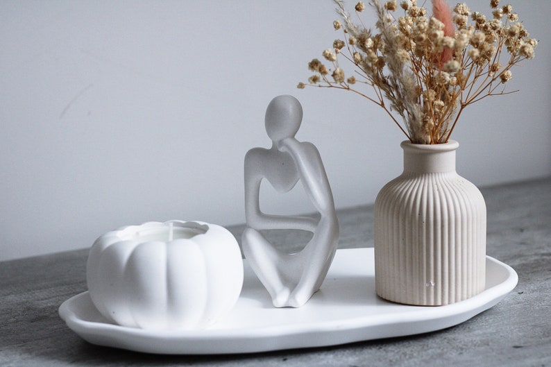 May include: White ceramic tray with a white pumpkin shaped candle, a white ceramic sculpture of a person sitting with their chin on their hand, and a white ceramic vase with dried flowers.