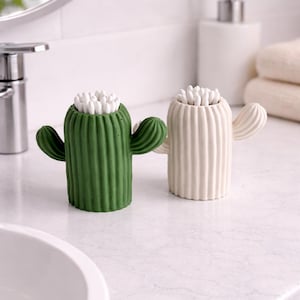 Può includere: Due porta cotton fioc a forma di cactus, uno verde e uno bianco sporco, pieni di cotton fioc bianchi. I porta cotton fioc hanno una consistenza a coste e sporgenze a forma di braccio. Sono su un piano bianco.