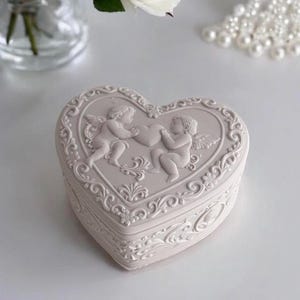 Könnte beinhalten: Eine herzförmige, cremefarbene Schmuckschatulle mit detailliertem Reliefdesign. Der Deckel zeigt zwei Engel, die ein Herz halten, umgeben von kunstvollen Schnörkeln. Die Box ist wahrscheinlich aus Keramik oder einem ähnlichen Material.