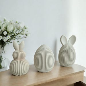 May include: Three off-white Easter decorations on a wooden surface. The decorations include two bunny figures and an egg-shaped figure. The bunny figures have vertical ribbed detailing and the egg-shaped figure has vertical ribbed detailing.
