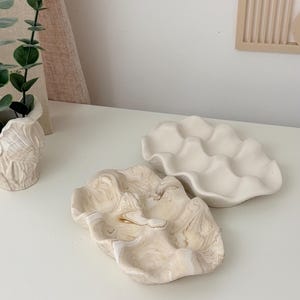 May include: Two decorative ceramic clam shell shaped dishes. One is a creamy white color with a wavy edge. The other has a marbled beige and white pattern. These dishes are ideal for holding jewelry or small items.