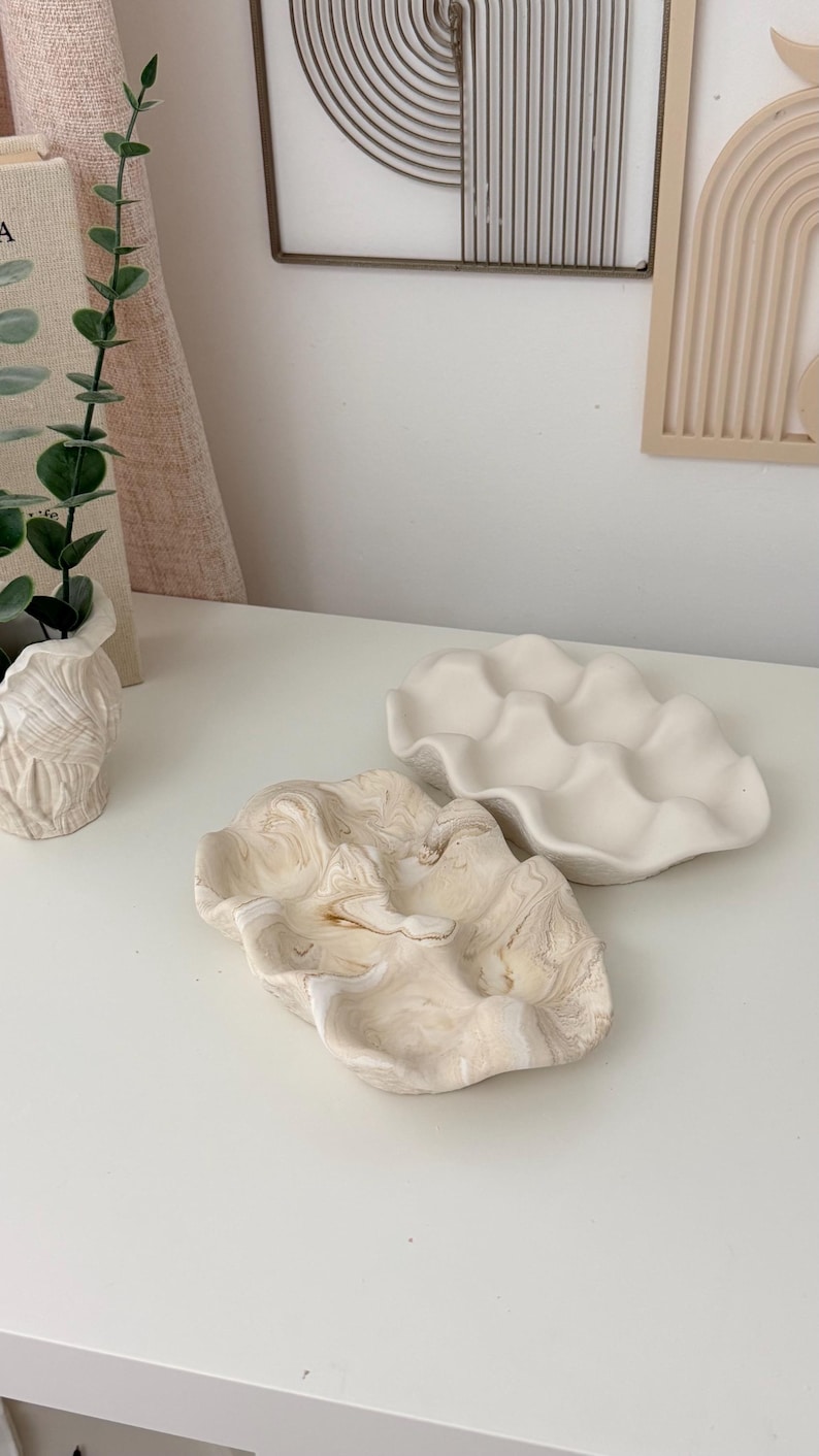 May include: Two decorative ceramic clam shell shaped dishes. One is a creamy white color with a wavy edge. The other has a marbled beige and white pattern. These dishes are ideal for holding jewelry or small items.