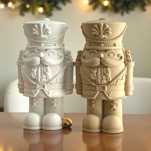 May include: Two decorative nutcracker figurines. One is off-white, the other is tan. Both have a traditional soldier design with a hat, mustache, and boots. The figurines are standing on a wooden surface, with a gold-coloured object between them.