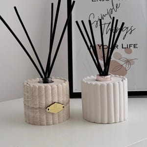 May include: Two reed diffusers with black reeds. One is beige with a gold label, the other is white. The background includes a framed print with the text "ENJOY THE SIMPLE THINGS IN YOUR LIFE."