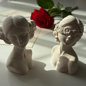 Puede incluir: Dos bustos de mujeres de yeso blanco con diademas florales. Un busto tiene una diadema que cubre los ojos. Los bustos est&aacute;n sobre una superficie blanca.