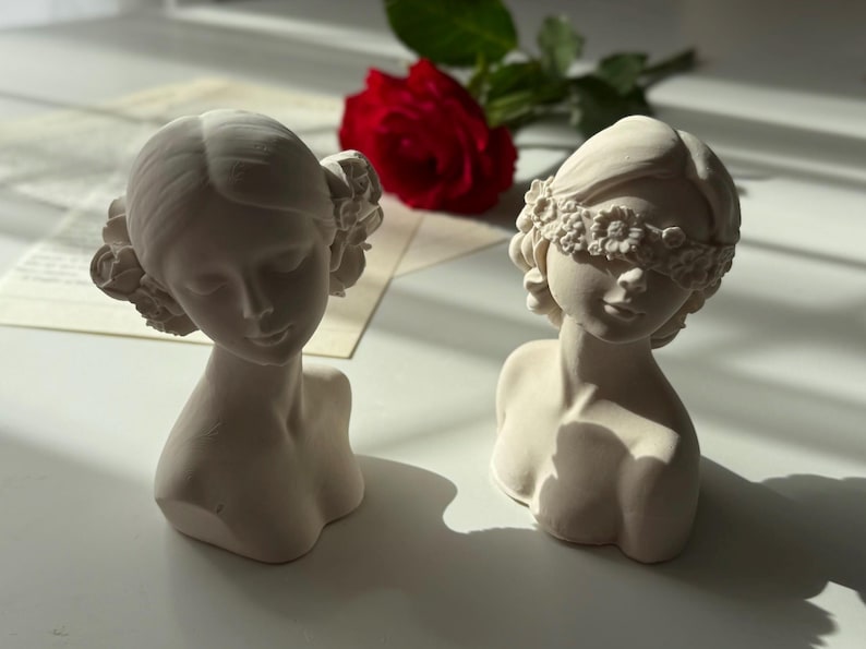 Puede incluir: Dos bustos de mujeres de yeso blanco con diademas florales. Un busto tiene una diadema que cubre los ojos. Los bustos est&aacute;n sobre una superficie blanca.