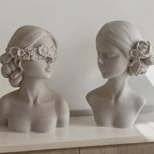 Puede incluir: Dos bustos de mujeres de yeso blanco con detalles florales. Un busto tiene una diadema floral que cubre los ojos, mientras que el otro tiene una sola flor detr&aacute;s de la oreja.