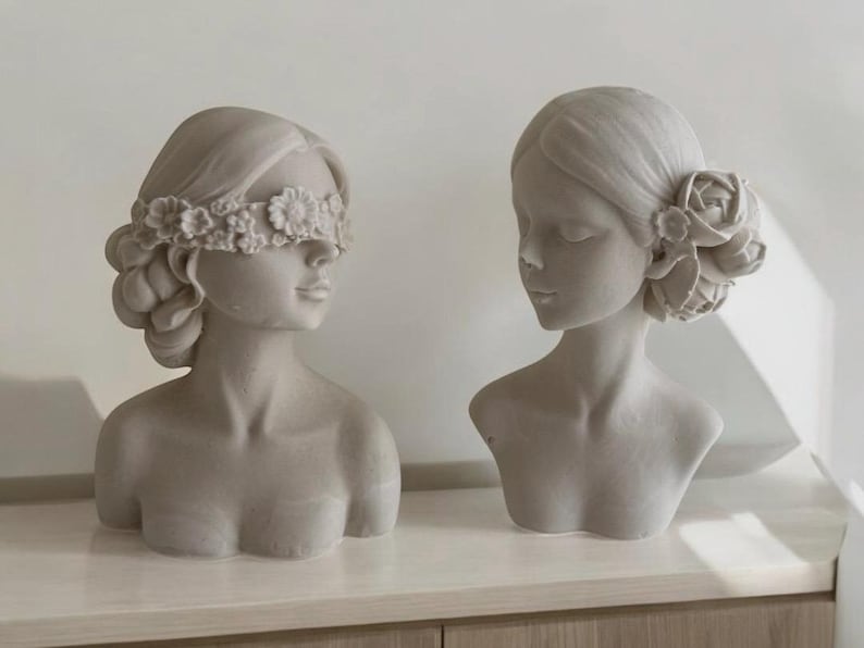 Puede incluir: Dos bustos de mujeres de yeso blanco con detalles florales. Un busto tiene una diadema floral que cubre los ojos, mientras que el otro tiene una sola flor detr&aacute;s de la oreja.
