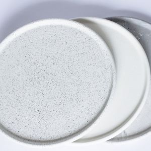 May include: Three round ceramic plates in different finishes. The first plate is white with black speckles, the second is plain white, and the third is white with gray marbling.