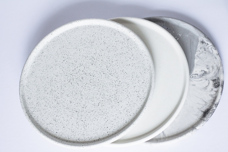 May include: Three round ceramic plates in different finishes. The first plate is white with black speckles, the second is plain white, and the third is white with gray marbling.