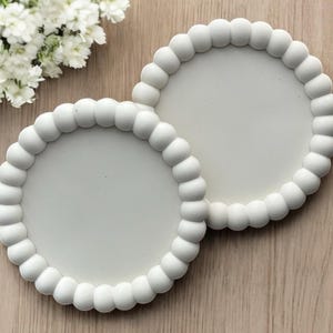 May include: Two white ceramic coasters with a raised, beaded edge. The coasters are round and have a smooth, flat surface.