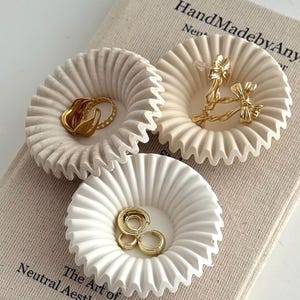 May include: Three white, ruffled, circular trinket dishes holding gold jewelry, including a ring and earrings. The dishes sit on a beige book with the text "HandMadebyAny" and "The Art of Neutral Aesthetic Decor".