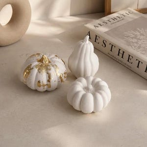 May include: Three decorative pumpkins in white. One pumpkin features gold leaf accents on its surface. The pumpkins vary in shape, with one being round and squat, one being taller and pear-shaped, and one being a smaller, round, ribbed pumpkin.
