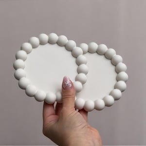 Pode incluir: Duas bases brancas e redondas com borda de contas. As bases são seguradas por uma mão com uma unha rosa clara com um desenho de laço branco. As bases estão em um fundo rosa claro.