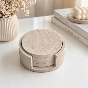May include: A set of circular coasters with a beige and white marbled pattern, stacked neatly inside a matching cylindrical holder with a front cutout. The coasters feature a smooth, stone-like finish with swirling, organic lines throughout.