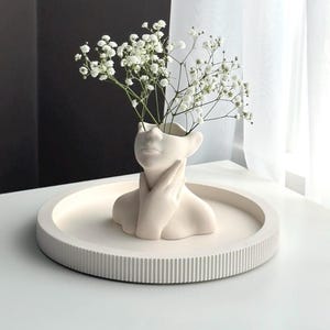 Puede incluir: Jarrón de cerámica blanca con forma de busto humano con las manos juntas delante del cuello. El jarrón está lleno de flores blancas de aliento de bebé y está sentado en una bandeja redonda blanca con un borde texturizado.