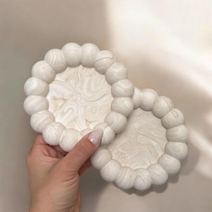 Pode incluir: Duas bases redondas brancas com um design marmorizado. Cada base tem uma borda com contas. As bases são feitas de um material sólido e são adequadas para segurar bebidas. Um acessório decorativo para casa.