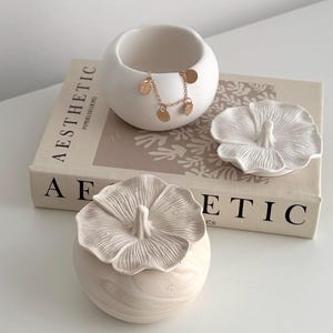 May include: Aesthetic home decor featuring a white ceramic bowl with a gold chain necklace and a small decorative flower. A book with the word "Aesthetic" is in the background. The items are on a white surface.