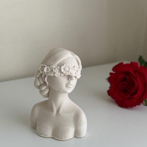 Puede incluir: Busto de mujer de yeso blanco con una corona de flores que le cubre los ojos. El busto est&aacute; sobre una superficie blanca junto a una rosa roja.