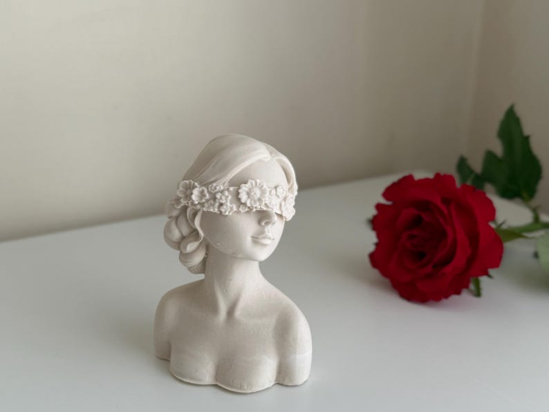 Puede incluir: Busto de mujer de yeso blanco con una corona de flores que le cubre los ojos. El busto est&aacute; sobre una superficie blanca junto a una rosa roja.