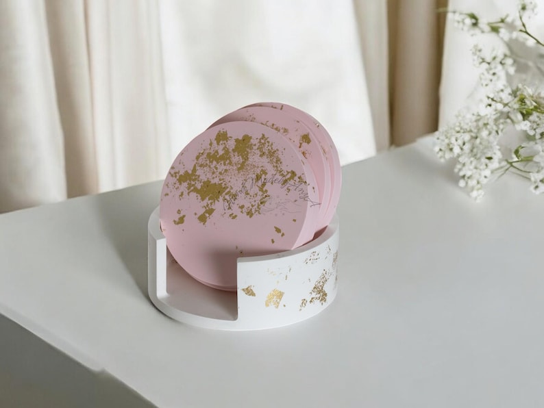 Puede incluir: Un juego de posavasos rosas con detalles de pan de oro, exhibidos en un soporte blanco. Los posavasos son redondos y tienen un dise&ntilde;o delicado y art&iacute;stico. El soporte tambi&eacute;n es blanco con detalles de pan de oro, creando un aspecto elegante.