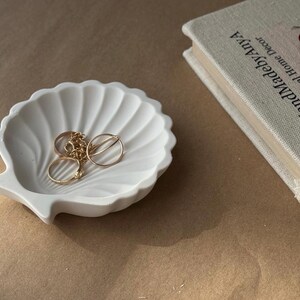 May include: White ceramic seashell dish with gold rings and a chain inside. The dish is on a brown surface.