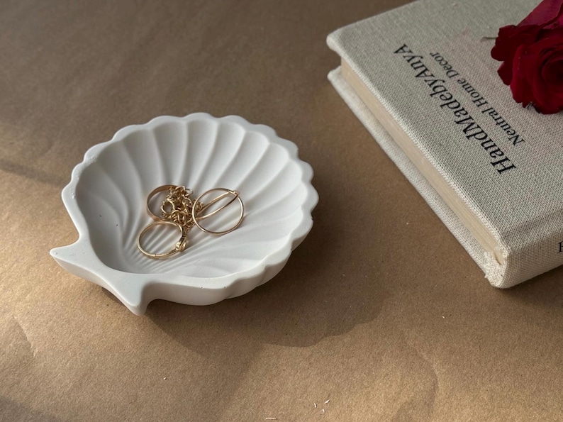 May include: White ceramic seashell dish with gold rings and a chain inside. The dish is on a brown surface.
