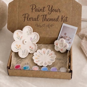 Puede incluir: Un kit de manualidades en una caja de cartón con la etiqueta Paint Your Floral Themed Kit que contiene tres objetos de cerámica blanca con forma de flor, detalles florales pintados, una pequeña fotografía y seis botes de pintura.