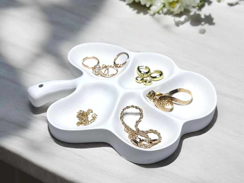May include: A white, leaf-shaped jewelry tray with multiple compartments. The tray holds various gold rings and necklaces. The tray has a handle on the side and sits on a light-colored surface.