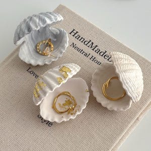 Puede incluir: Tres conchas marinas en forma de cuenco para joyas, cada una con una pieza de joyería dorada. Las conchas son blancas y grises, con superficies texturizadas. La joyería incluye un par de pendientes y dos anillos. Las conchas descansan sobre un libro.
