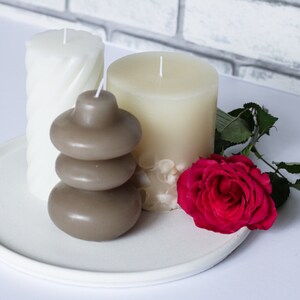 May include: Three candles on a white plate with a single red rose. The candles are white and brown, and the brown candle is stacked in three tiers.