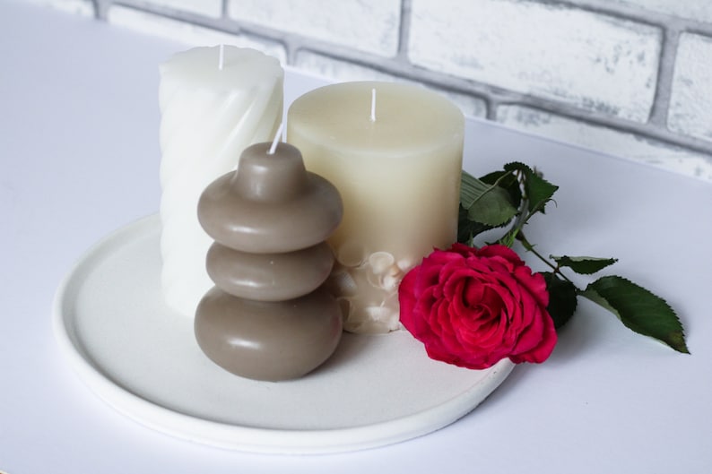 May include: Three candles on a white plate with a single red rose. The candles are white and brown, and the brown candle is stacked in three tiers.