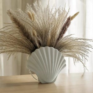 May include: A white ceramic seashell vase with a bouquet of dried pampas grass.