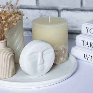 May include: A white tray with a white candle, a white vase with dried flowers, and a white decorative sculpture with a face. The tray is on a white surface. The candle is surrounded by white seashells. The books in the background have the text "Your So", "Take Time", and "What Ma".