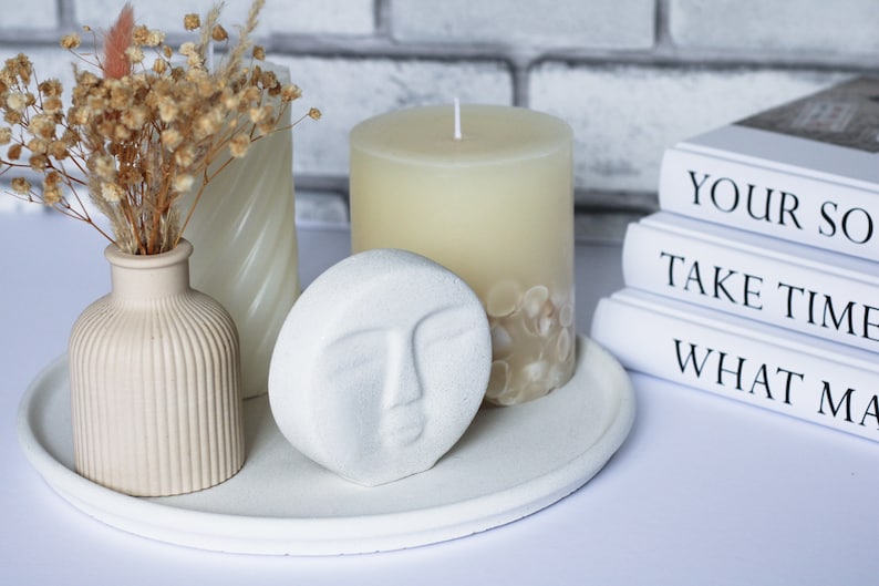May include: A white tray with a white candle, a white vase with dried flowers, and a white decorative sculpture with a face. The tray is on a white surface. The candle is surrounded by white seashells. The books in the background have the text "Your So", "Take Time", and "What Ma".