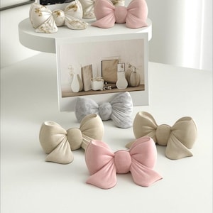 May include: Several decorative bow ornaments in shades of pink, beige, and marbled patterns. The bows are displayed on a white surface and a shelf, with a framed photograph visible in the background. The bows are made of a soft material.