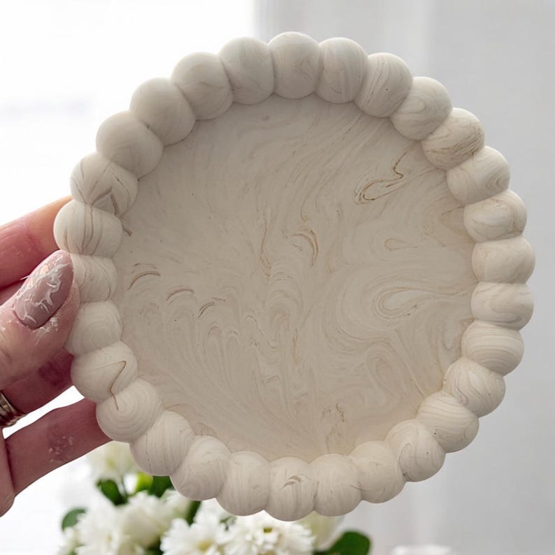 May include: A round, off-white decorative tray with a beaded edge. The tray has a marbled pattern in shades of beige and cream. The edge is made of small, rounded beads. The tray is a home decor item.