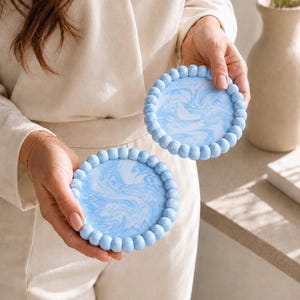 May include: Two circular coasters featuring a light blue and white marbled pattern on the flat surface. Each coaster is framed by a raised border composed of small, uniform, light blue spherical beads arranged in a continuous ring around the edge.