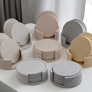 Puede incluir: Colecci&oacute;n de posavasos redondos en varios colores neutros, incluyendo beige, gris y blanco moteado. Cada juego de posavasos se muestra en un soporte a juego. Los posavasos est&aacute;n hechos de un material s&oacute;lido.