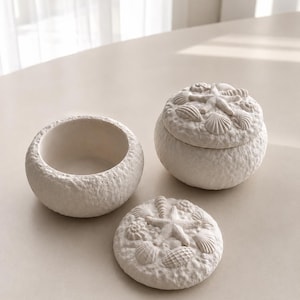 May include: Three off-white ceramic decorative pieces. One is a small, open bowl. Another is a round box with a lid, and the lid is decorated with seashells and a starfish. The third piece is the lid of the box.