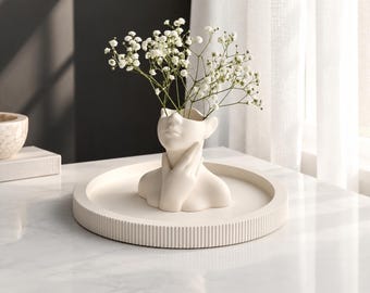May include: A matte off-white ceramic vase shaped like a human head and shoulders with hands touching the neck, holding white baby's breath flowers. The vase sits centered on a matching circular tray with a ribbed edge and a flat base.